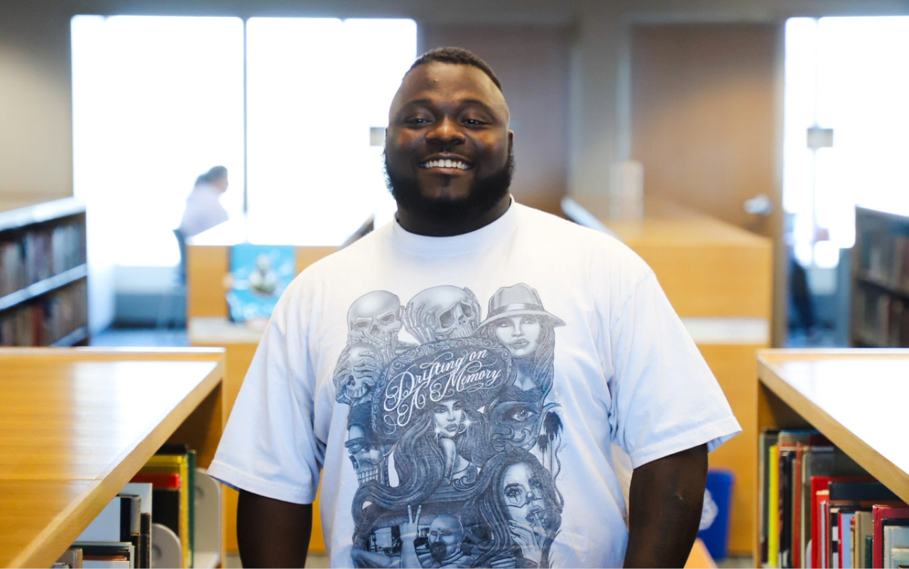 COSI student smiling at the camera in a library with bookshelves on either side